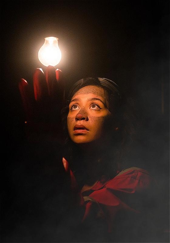 Close-up of a performer in darkness, looking up at a lightbulb
