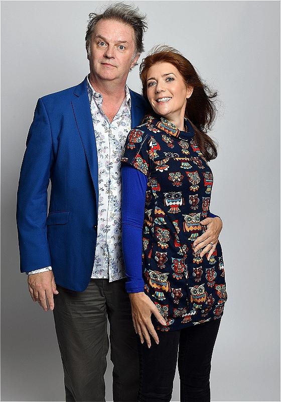 Paul Merton stand on the left wearing a blue suit. He has his around the waist of Suzi Webster, who wears a black patterned dress. 