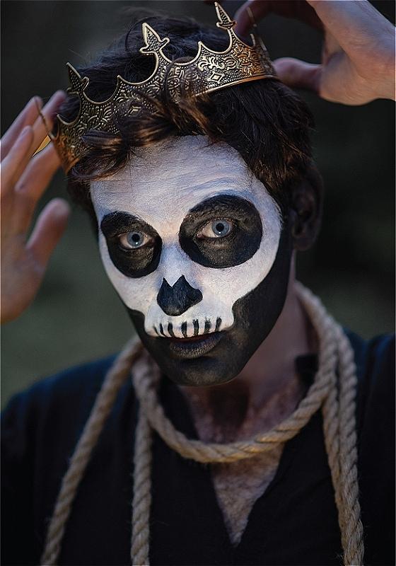Close-up of a man with his face painted like a skeleton, wearing a rope around his neck and a crown on his head.