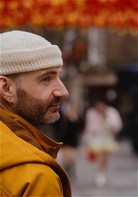 A side profile image of a performer, wearing a cream beanie hat and yellow coat. The background is blurred, with the silhouette of people walking down the street. 