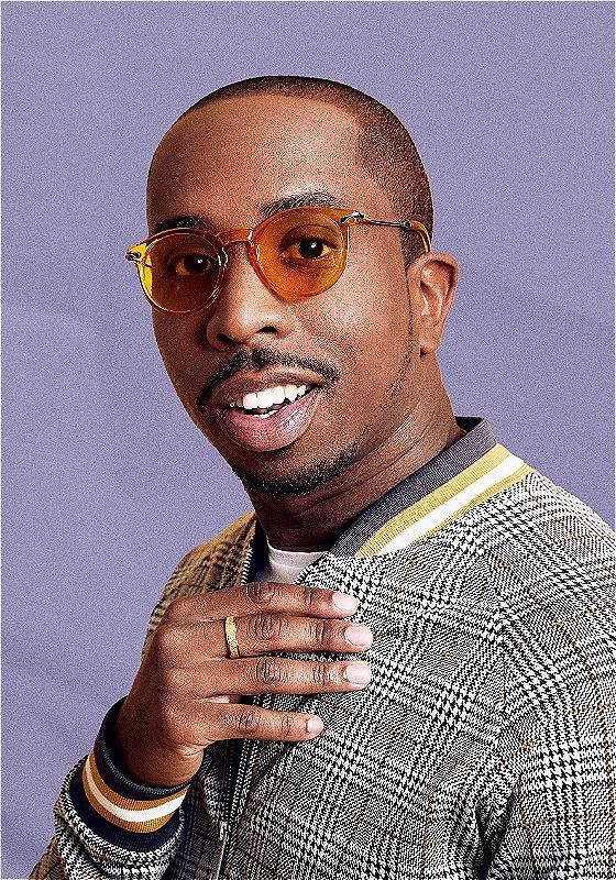 The performer looks into the camera, wearing a check jacket and sunglasses. They are in front of a purple background. 