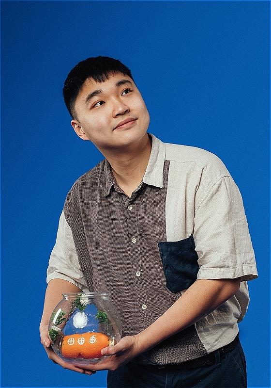 The performer in front of a blue background, holding a fish bowl with a toy submarine inside. 