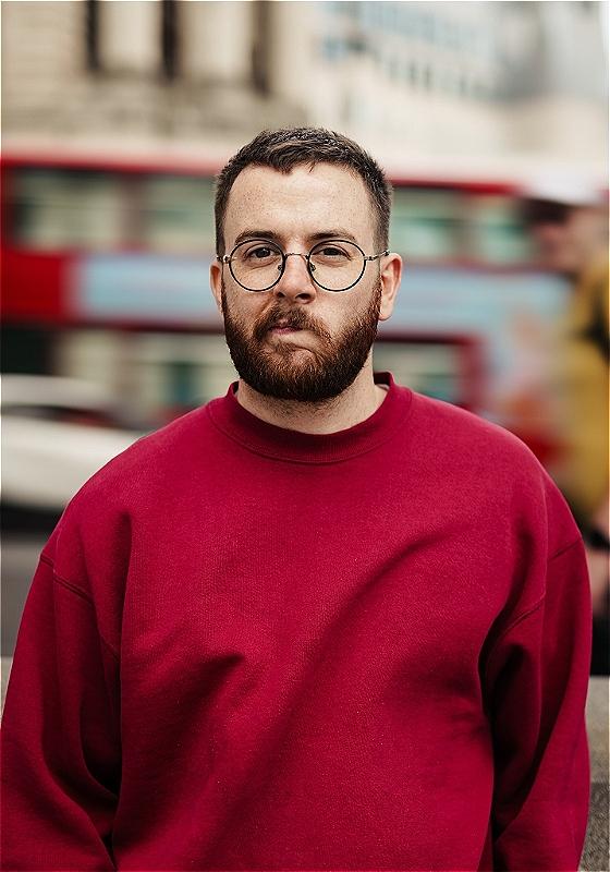 Rich looks directly at the camera wearing a red jumper as a London bus passes in the background.