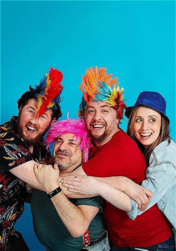 Four performers wearing colourful spiky wigs embrace with big smiles