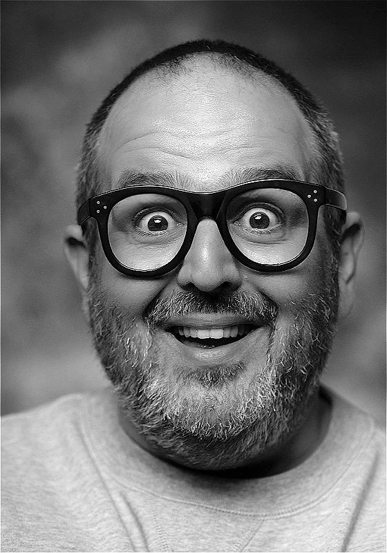 A black and white image of the performer's face. He wears glasses and his eyes are wide. 