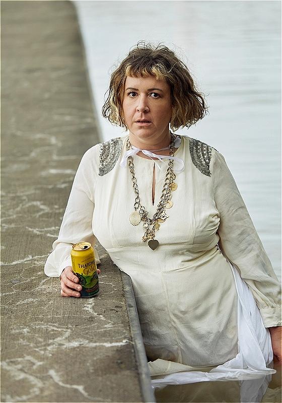 Amy standing in a pond, wearing a long white dress and holding a can of cider.