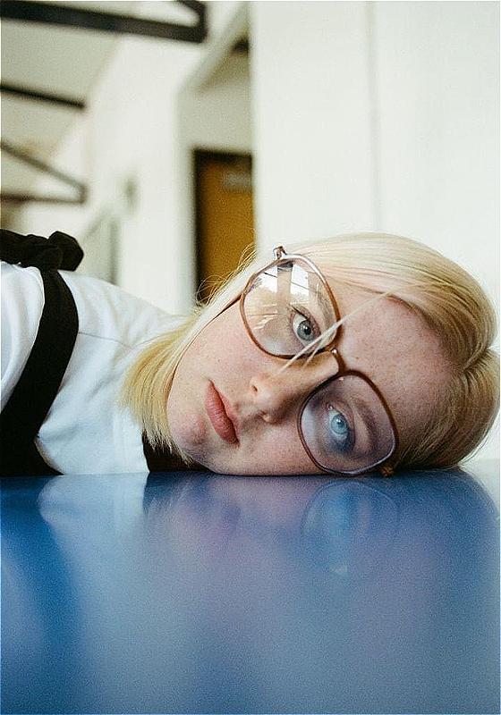 A performer wears glasses and has their head on a blue table, looking into the camera with a straight face.
