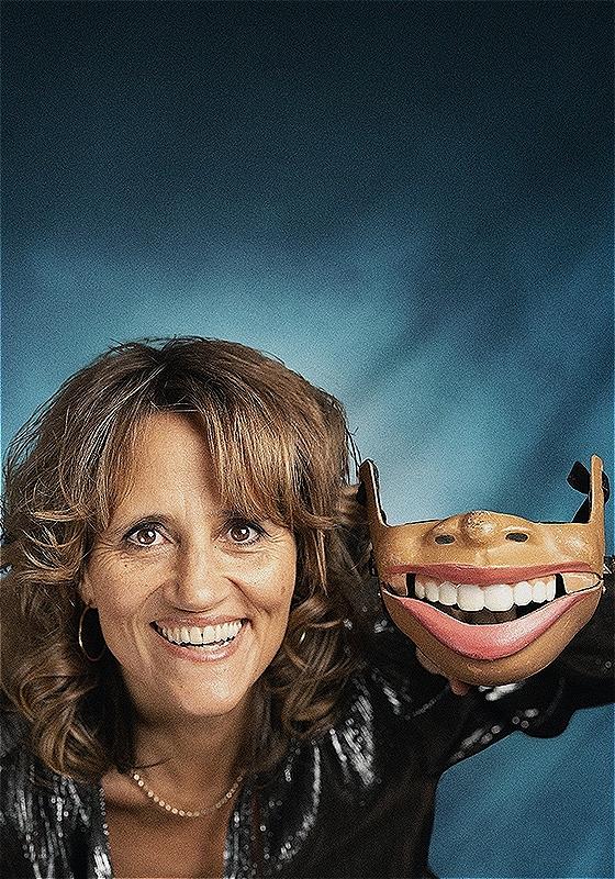 The performer looks into the camera smiling. In her right hand is a puppet that consists of a nose and mouth.