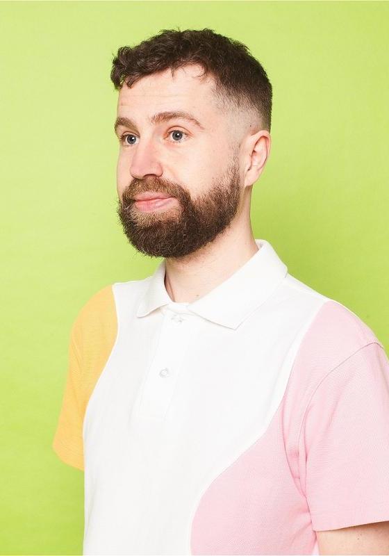 The performer is in front of a bright green background, wearing a white polo shirt with pink and yellow on the sleeves.