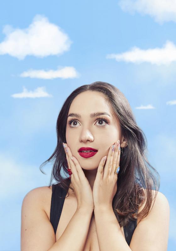 In front of a hazy blue sky backdrop, Bella stands with hands gently cupping their chin. Performer wears bright red lipstick.