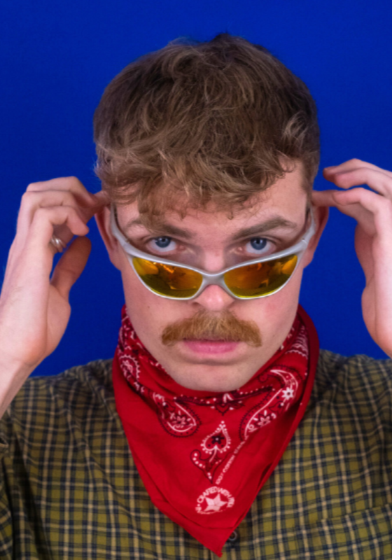 Horatio squares up to a blue background wearing a yellow checked shirt, red bandana around their neck and holding a pair of retro sunglasses halfway down their nose.