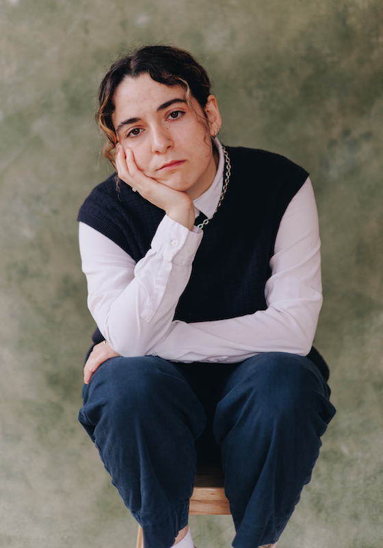 Rachel wears a navy blue tank top over a white shirt and blue trousers. They sit on a stool with one arm draped over their knees and the other which their chin is resting on.