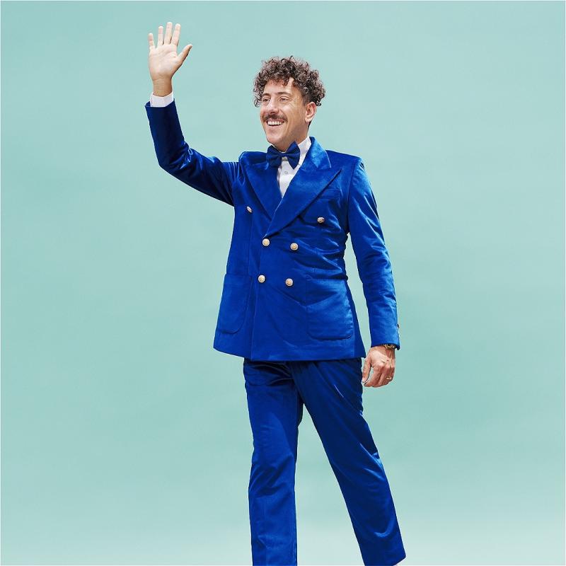 On a turquoise background, the performer is wearing a bright blue suit, smiling and waving their right hand. 