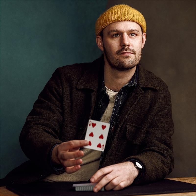 Andrew sits at a table, looking off camera and holding the six of hearts.