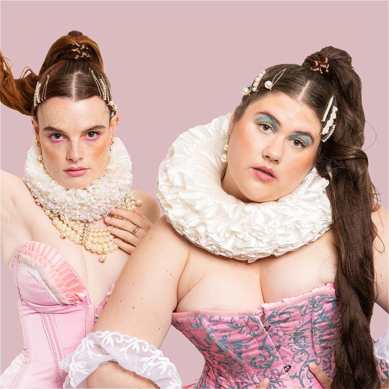 Two performers in front of a pink background wearing pink corsets and white collars. Both have ponytails and colourful eyeshadow.
