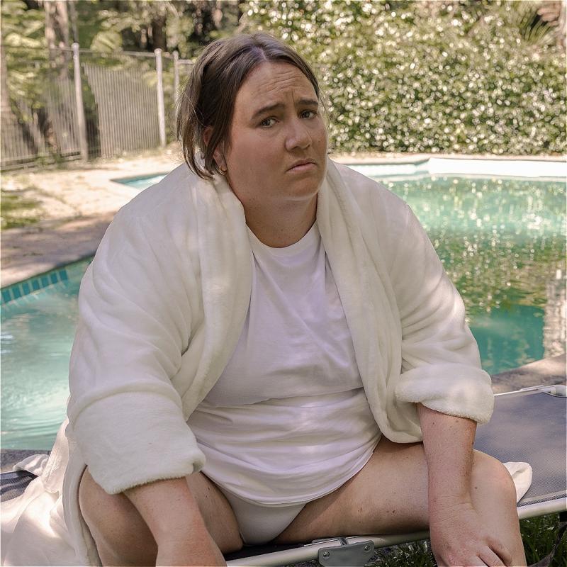 The performer sits in a garden in front of a swimming pool, wearing a white dressing gown.