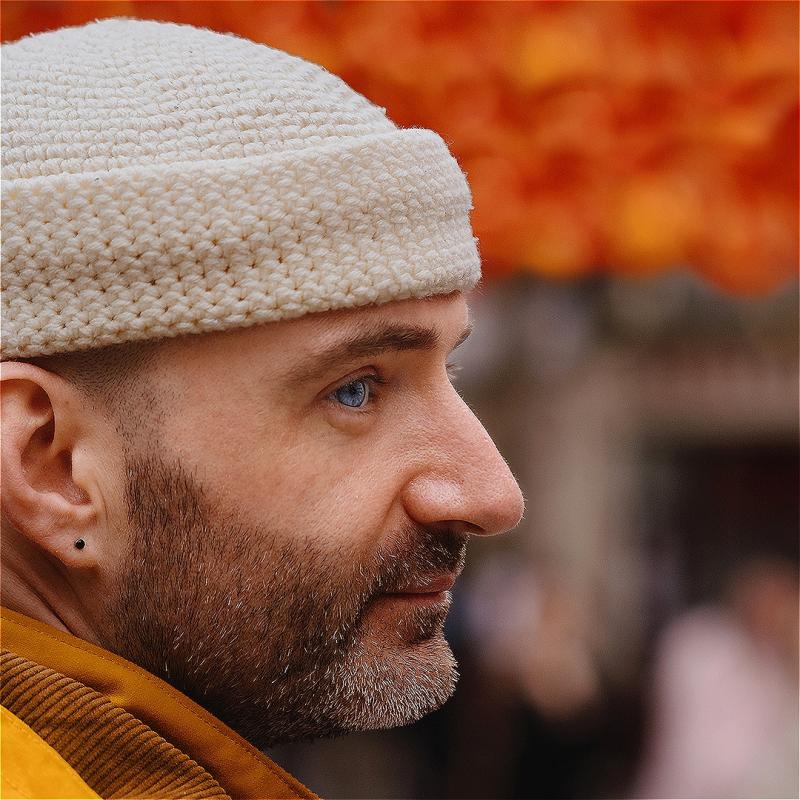 A side profile image of a performer, wearing a cream beanie hat. The background is blurred.