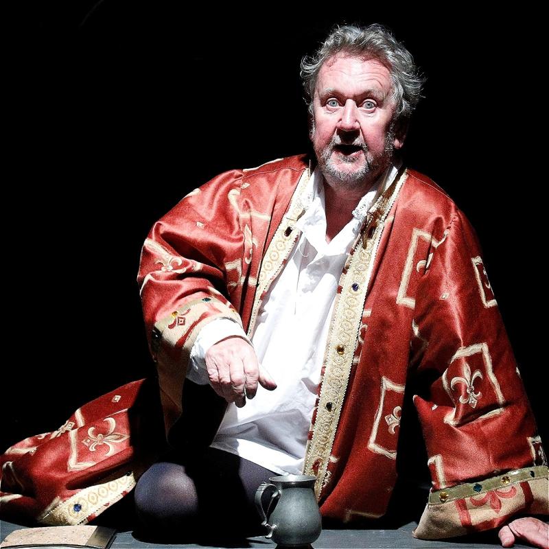 A man sits down onstage wearing a red printed silk robe