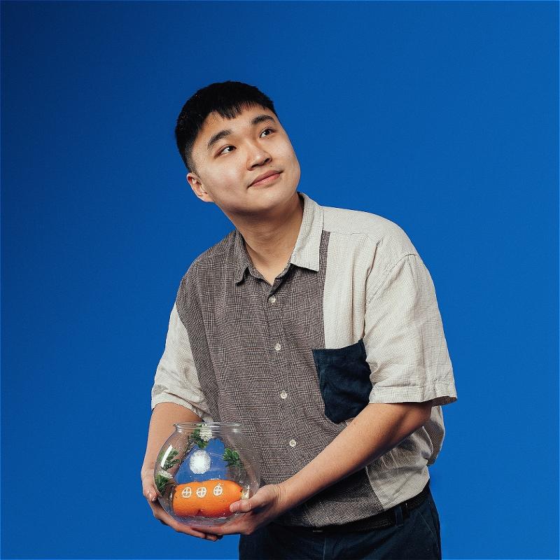 The performer in front of a blue background, holding a fish bowl with a toy submarine inside. 