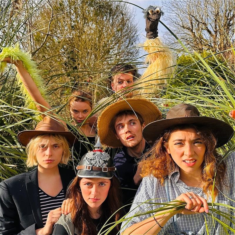 Five performers wear adventure hats amongst some tall grass. Behind them is a puppet emu. 