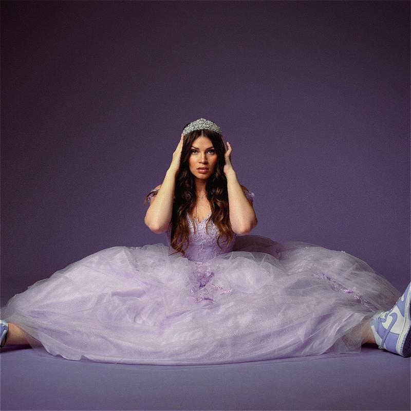Katie sits on the floor in a poufy purple dress and tiara with a matching background