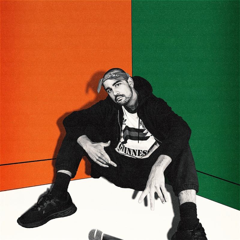 The performer sits in a corner in black and white. The walls behind him painted green, orange and white to represent the Indian flag. A microphone lies on the floor.