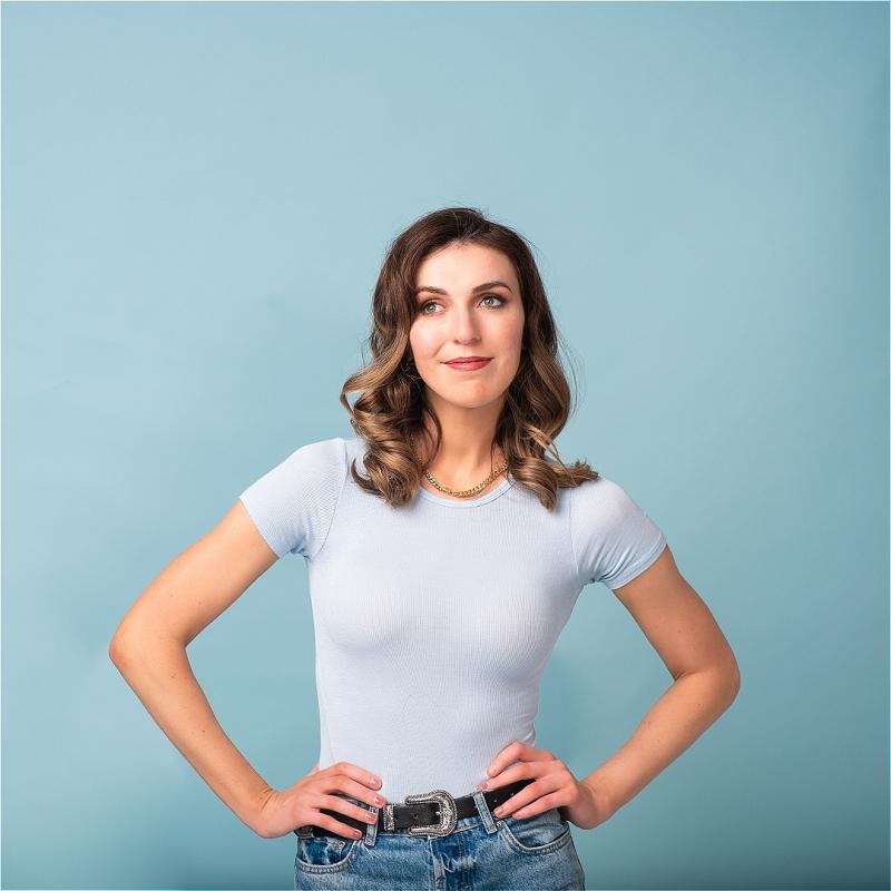 Melanie stands against a pale blue wall with her hands on her hips.