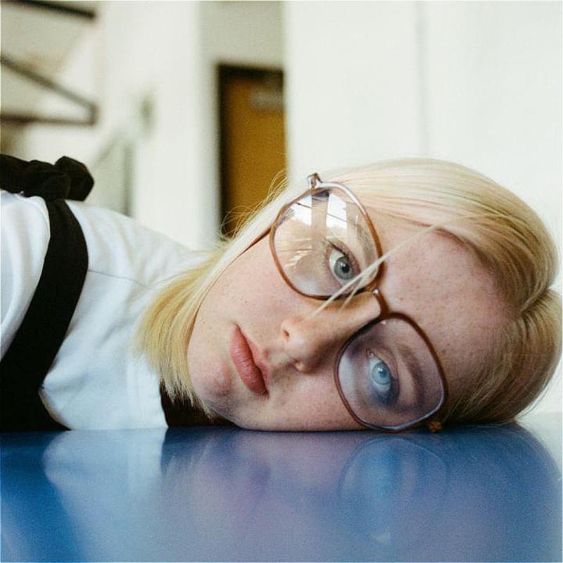 A performer wears glasses and has their head on a blue table, looking into the camera with a straight face.