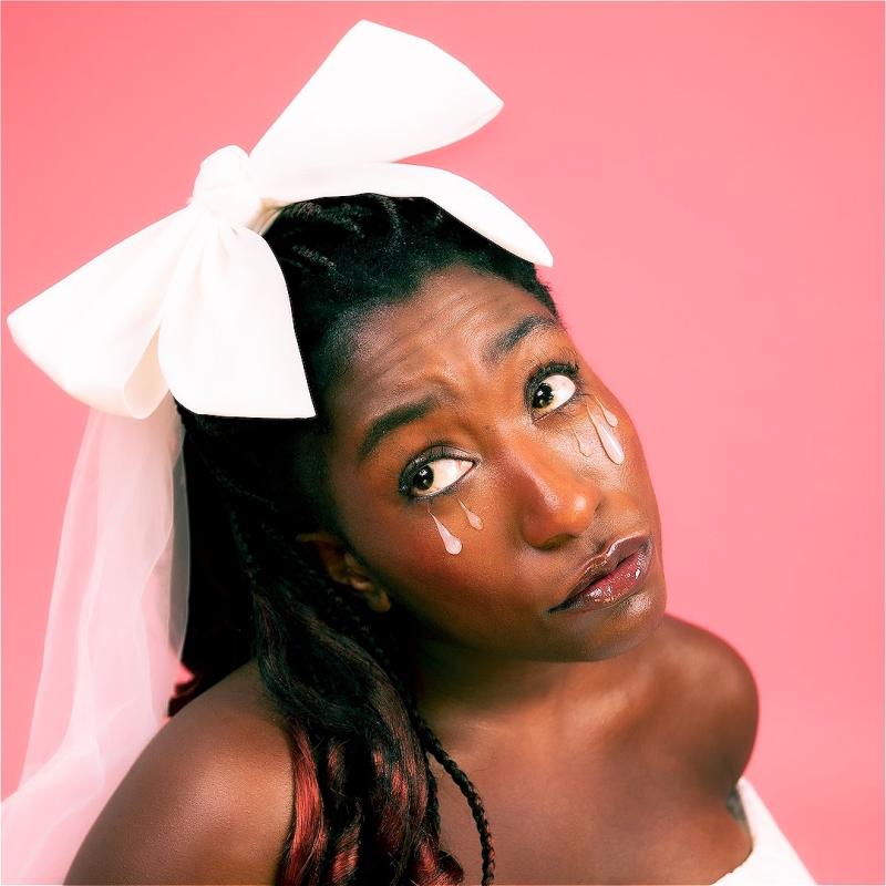 The performer is in front of a pink background with a white bow in her hair and white teardrops drawn on her face