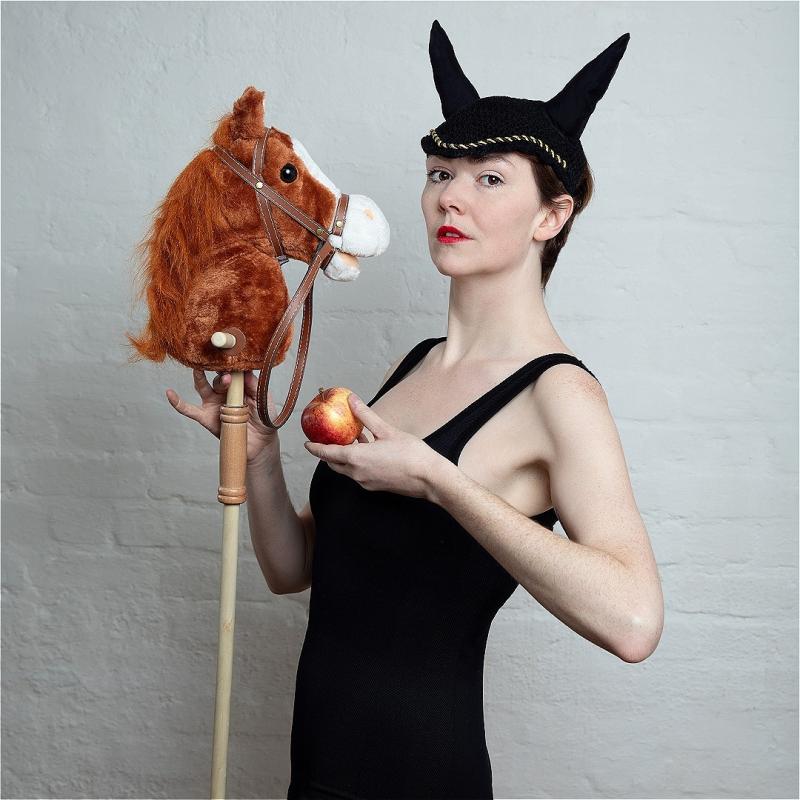 The performer wears black horns and looks to the camera holding an apple and a toy horse.