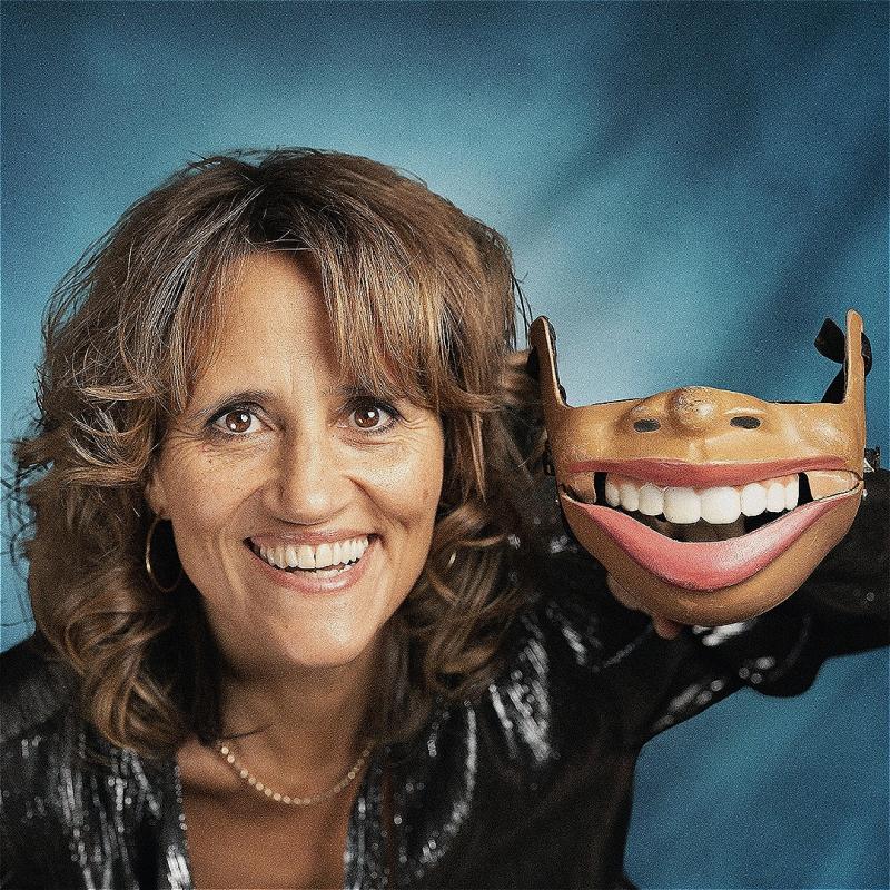 The performer looks into the camera smiling. In her right hand is a puppet that consists of a nose and mouth.