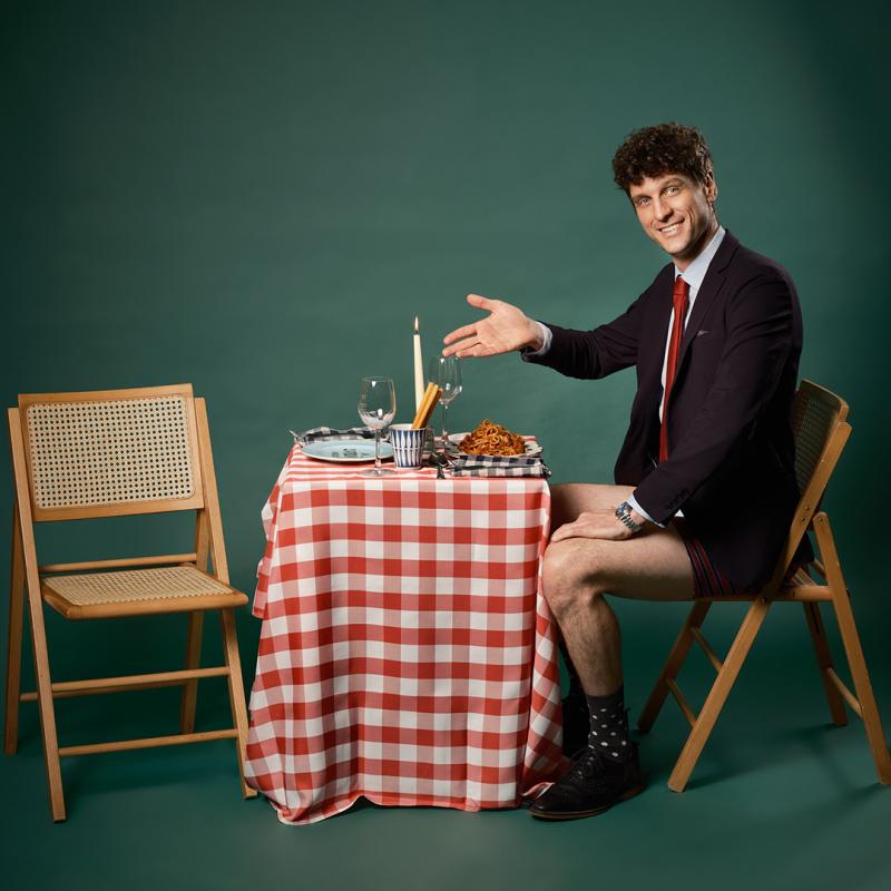 The performer sits at a table with plates and wine glasses. He wears a black suit and underpants, smiling and gesturing to the empty seat opposite. 