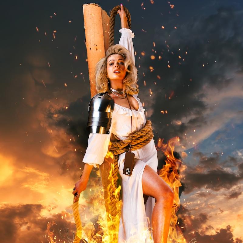 Abi wearing white shirt and shoulder armour is tied to a plank of wood with sparks of fire surrounding her