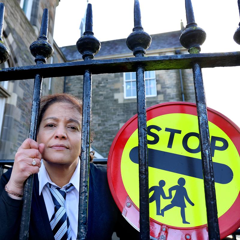 The performer peers through the railing on a school playground, wearing a school uniform and holding a stop sign.
