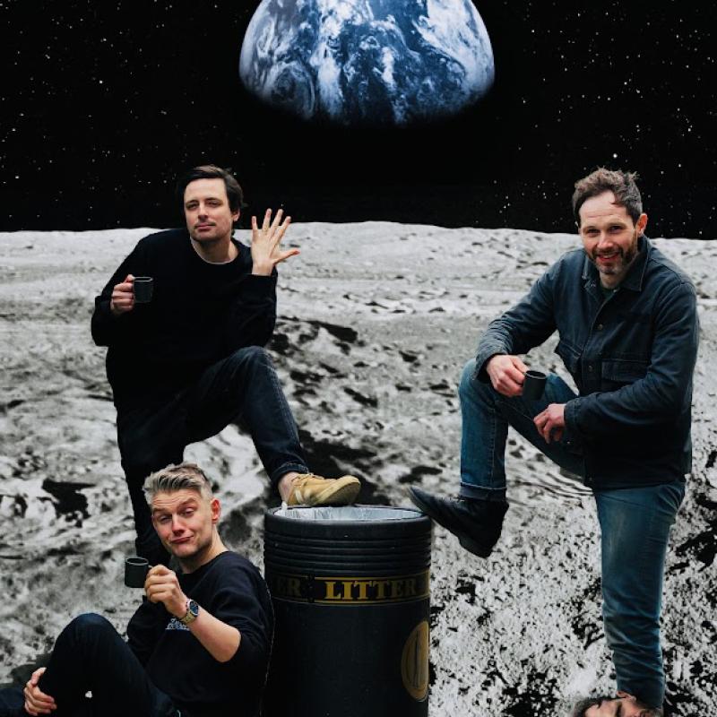 Three performers in front of a simulated background displaying a photograph of earth as seen from the surface of the moon. There's a bin in between the three performers. One of the performers has a foot propped on the bin, and their other foot is a face. The second performer also has a foot propped up on the bin, is holding up a hand, and their other hand is holding a mug. The third performer is leaning with their back to the bin, and is holding up a mug in a toast to the camera. 